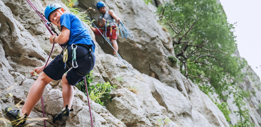 Smiling teen boy in protective helmet abseiling from cliff rock wall using rope